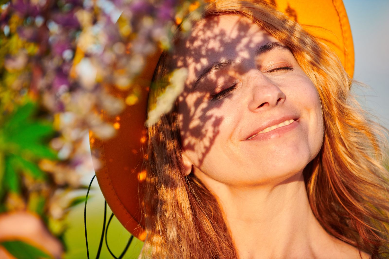 Smiling woman with closed eyes in an orange sun hat, leaf shadows across her face in sunlight