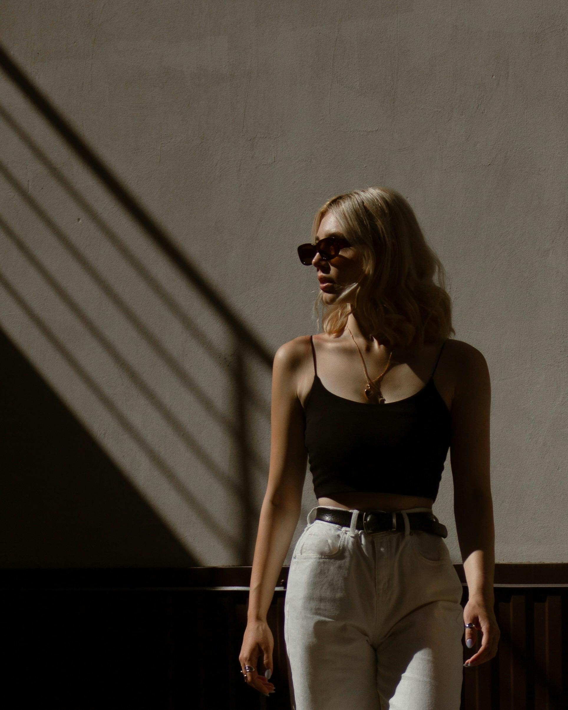 Woman with sunglasses and blonde hair stands in dramatic lighting, wearing a black tank top.
