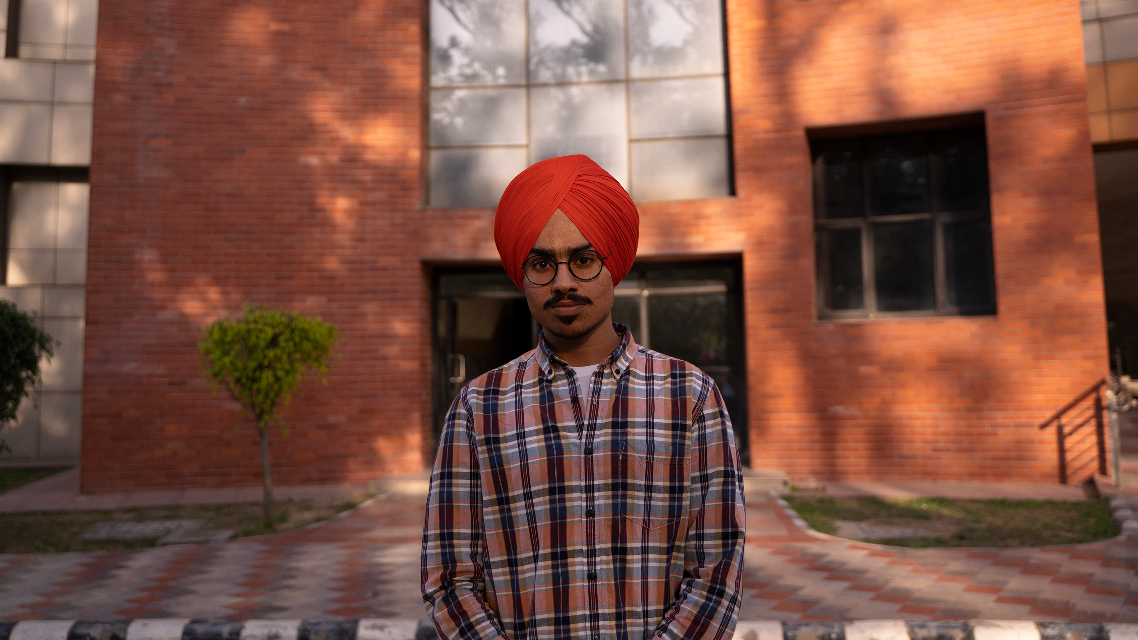 A man with a red turban and plaid shirt stands confidently in front of a brick building.