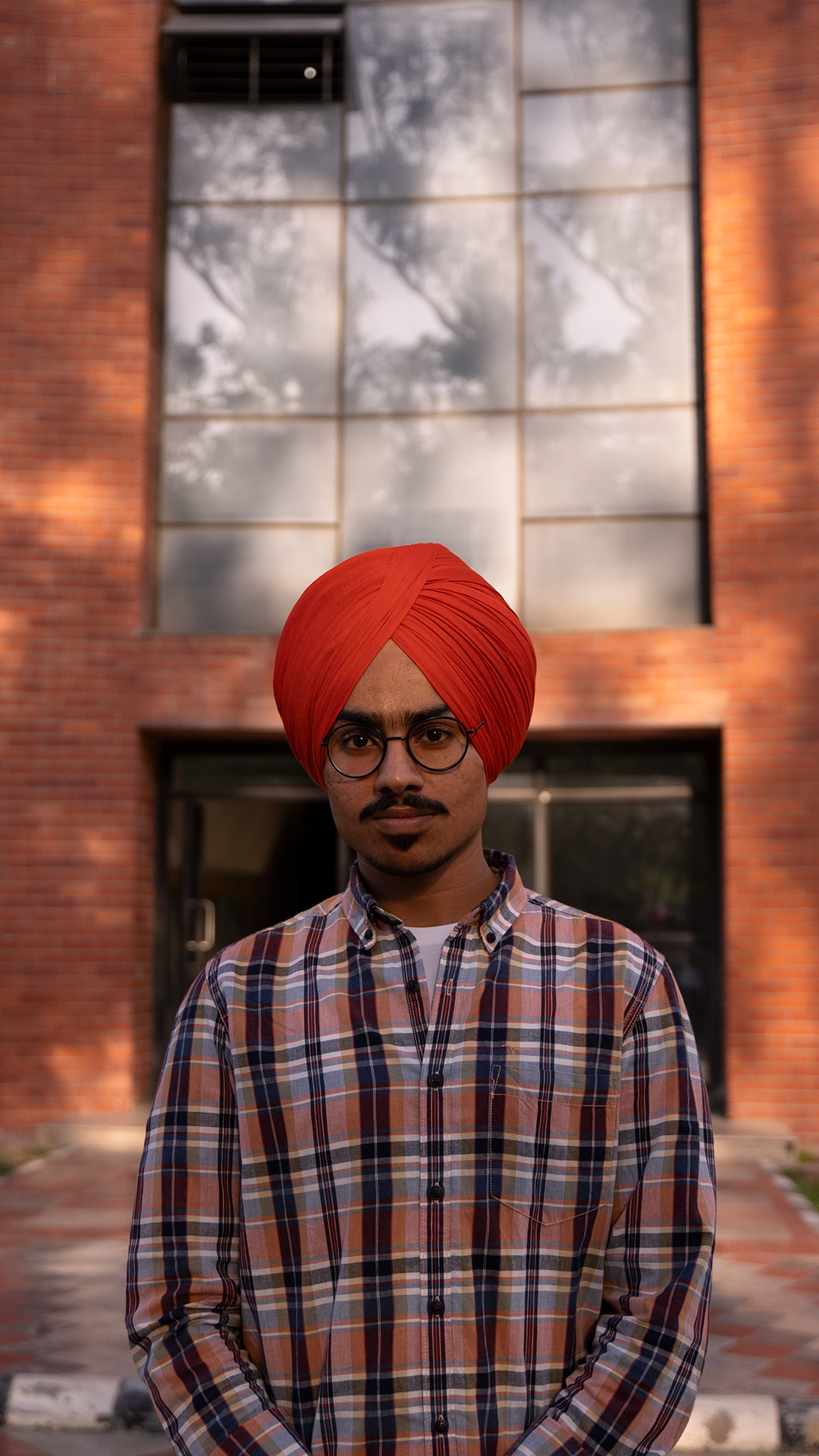 Portrait of a young man wearing glasses and a turban, standing outdoors against a brick building.