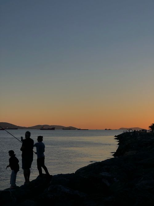 Free A family enjoys fishing at sunset, casting silhouettes against a colorful sky over a tranquil lake. Stock Photo