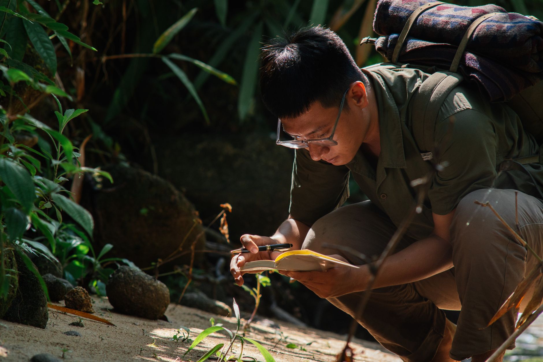 Man crouching outdoors, writing in a notebook surrounded by lush greenery, showcasing a peaceful, contemplative moment.