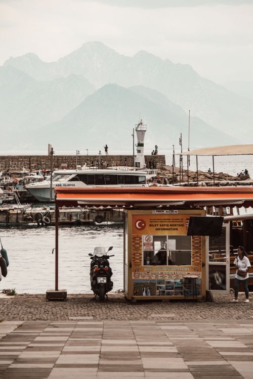 Free A peaceful harbor with a lighthouse, distant mountains, boats, and a small kiosk on a cloudy day. Stock Photo