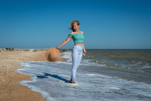 A Woman in Teal Tank Top and Blue Pants Holding a Brown Hat Standing on the Beach