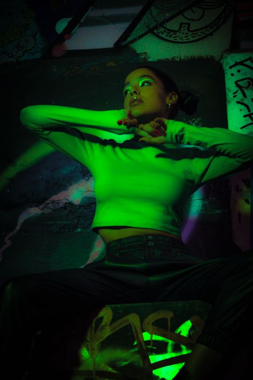 Free A young woman poses boldly against a graffiti-covered wall in an urban setting with dynamic green lighting. Stock Photo