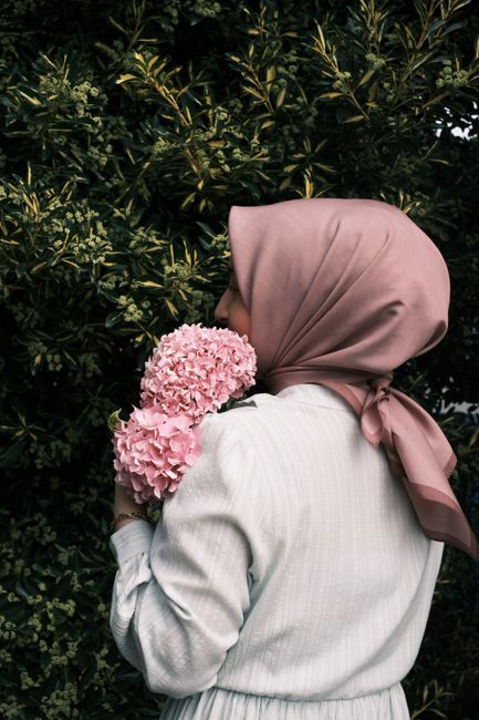 Woman in hijab enjoying pink hydrangea flowers against lush greenery outdoors.
