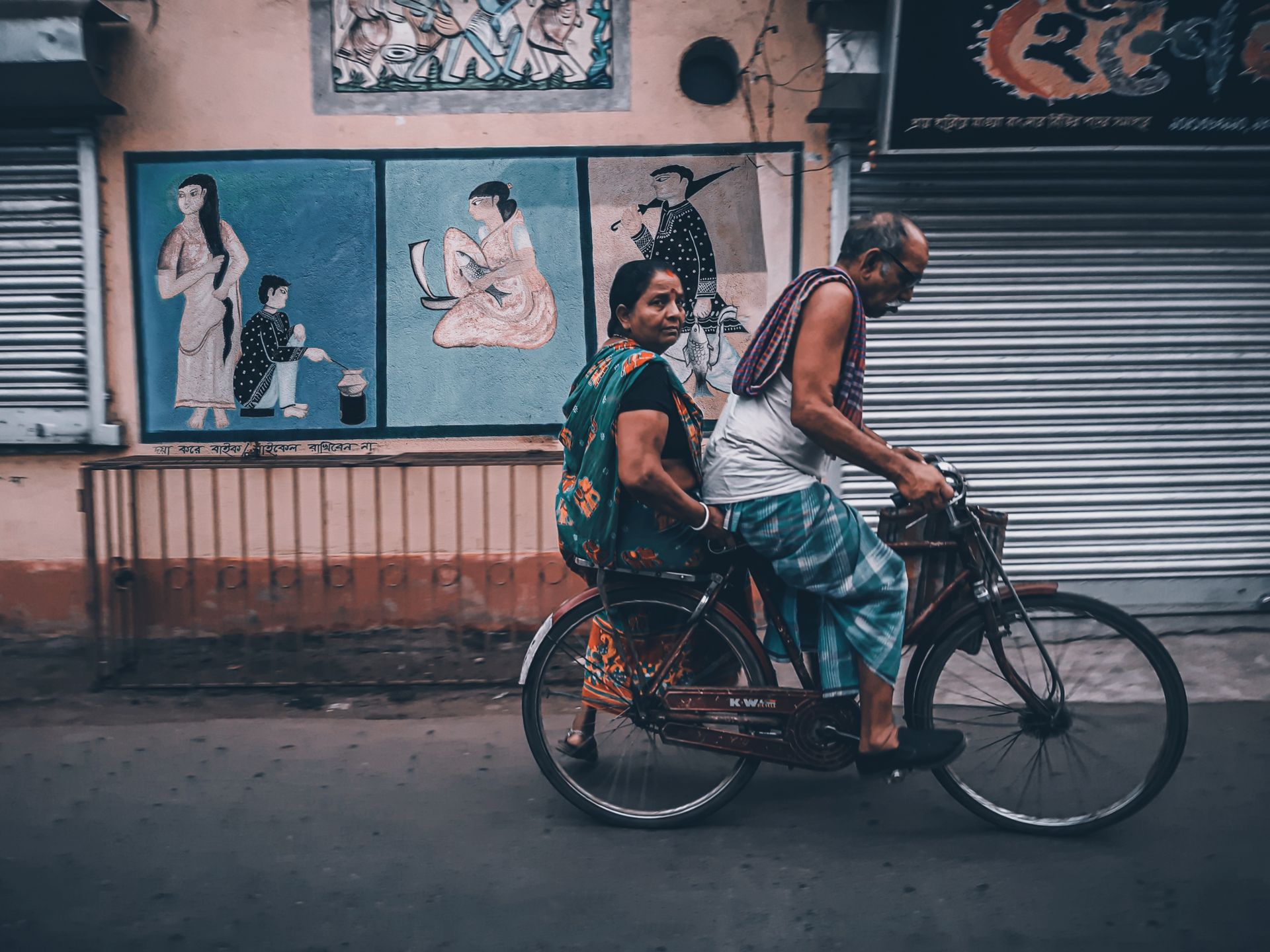 A couple rides a bicycle past vibrant murals in Kolkata, India, showcasing daily life in an urban setting.