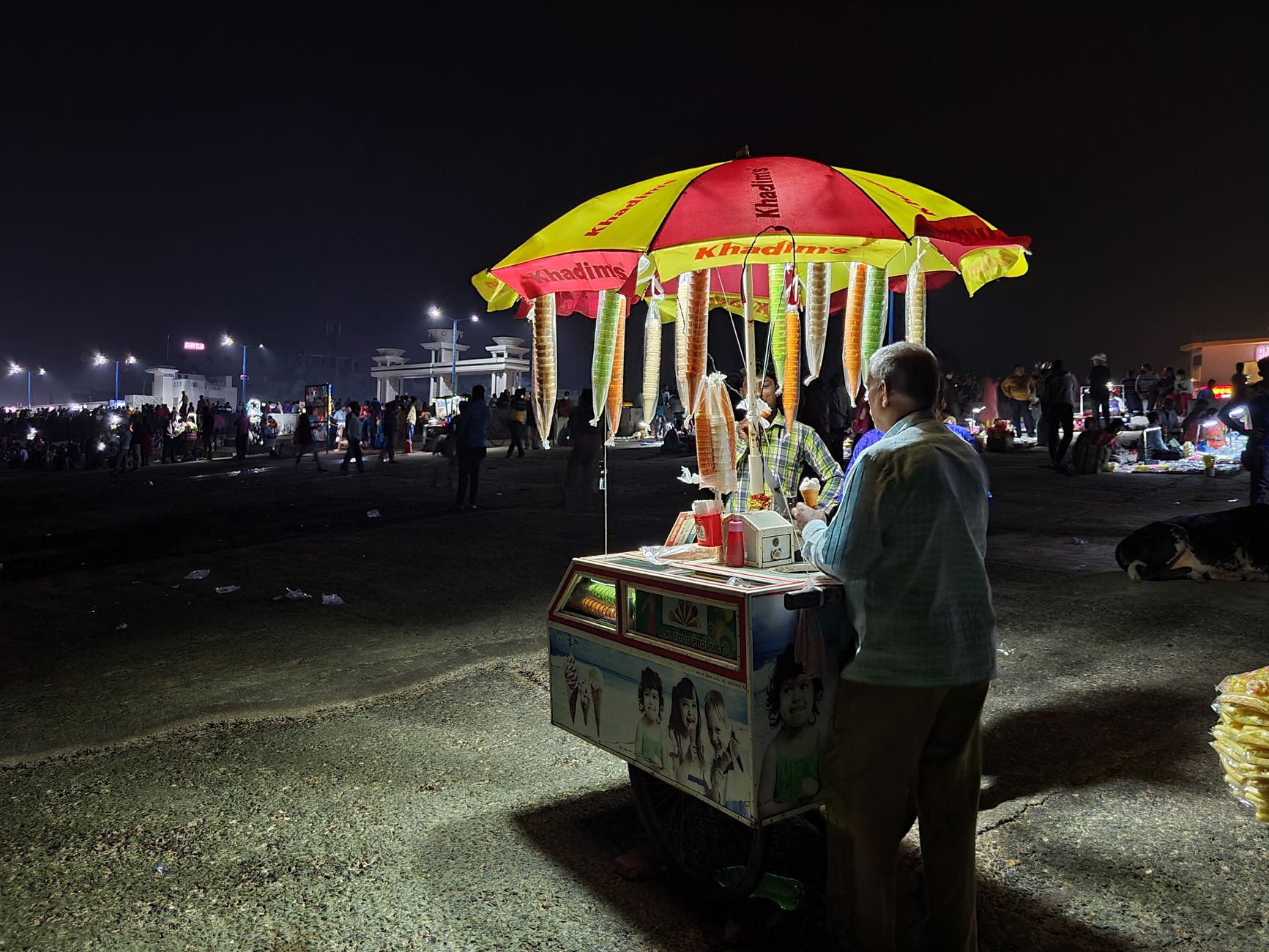 A street vendor sells ice cream cones at night in Digha, India, under a vibrant umbrella.