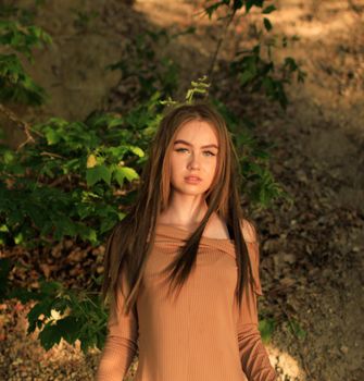 A serene portrait of a young woman with long brown hair posing outdoors amidst green leaves and natural light.