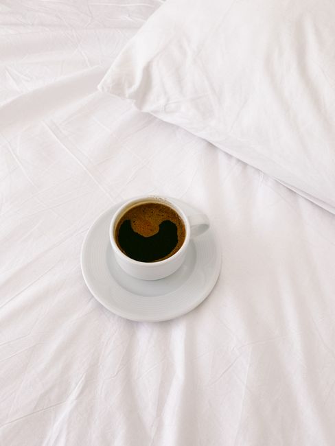 A cup of black coffee on white bed linens symbolizes a relaxing morning.