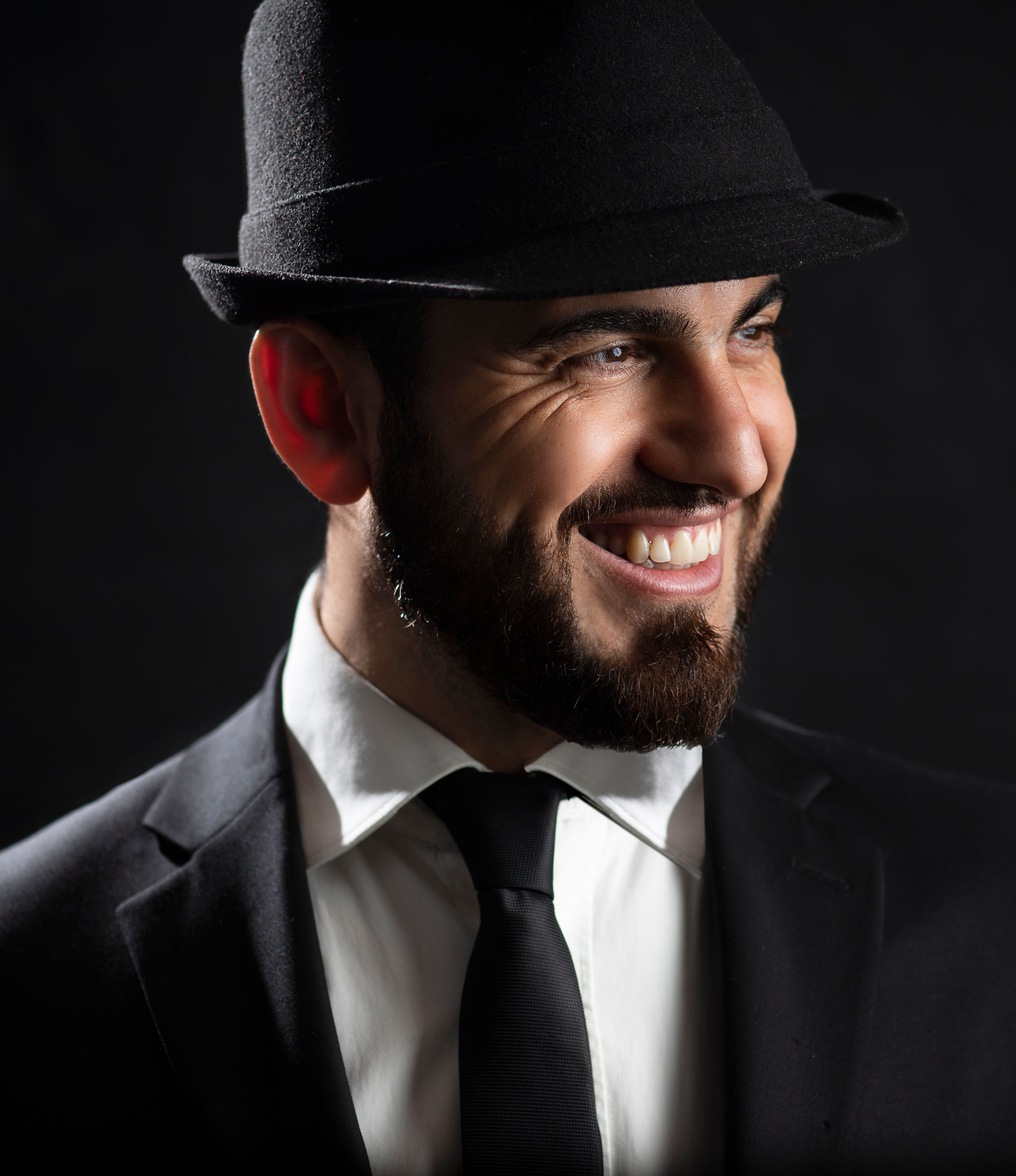 Stylish bearded man in a black hat and suit posing with a smile against a studio backdrop.