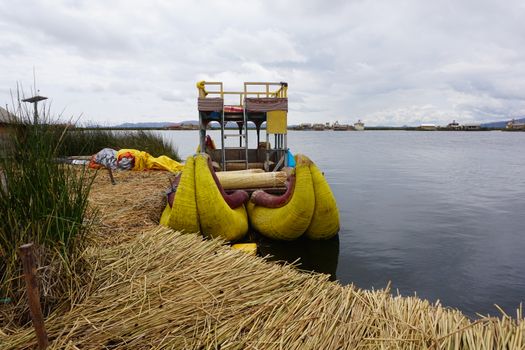 A traditional reed boat docked on a reed-covered shore of a calm lake under a cloudy sky.