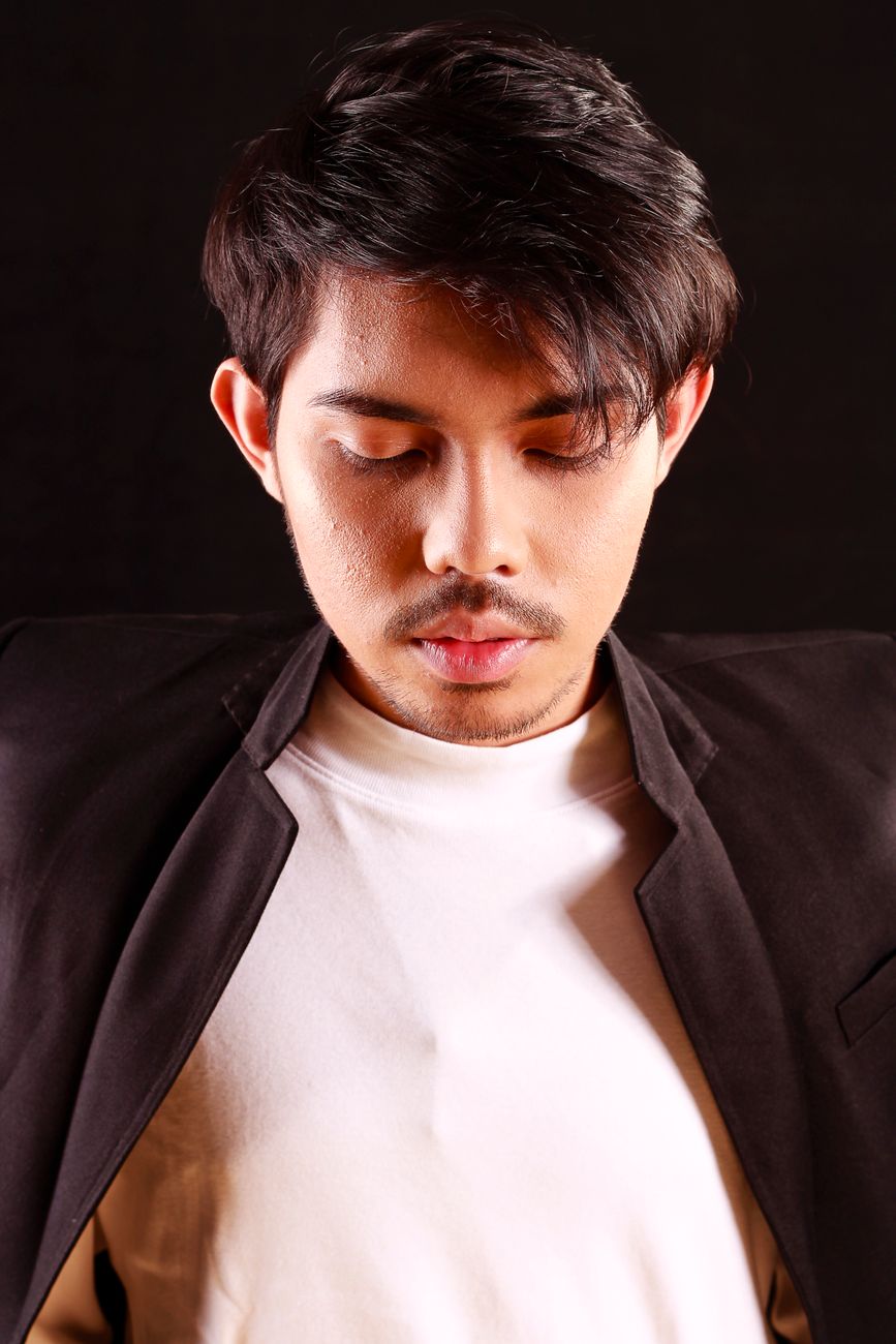 A serious young man in a black coat looking down, captured in a studio setting.