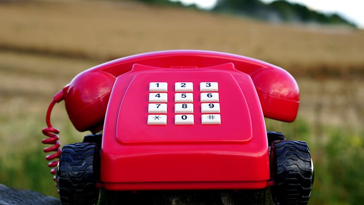 Red Rotary Phone With Black Wheels Near Brown Grasses During Day Time