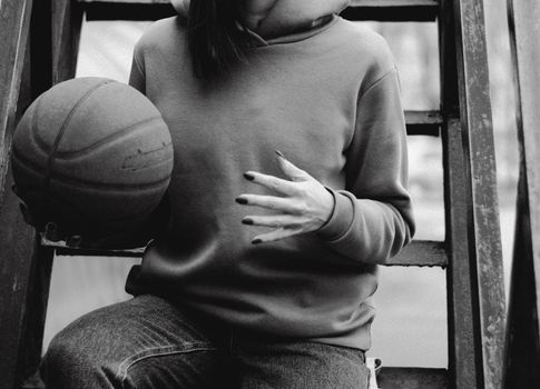 Black-and-white image of a person in hoodie holding a basketball while sitting on a metal ladder.