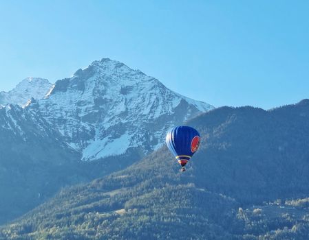 A vibrant hot air balloon floats over the snow-capped Italian Alps in clear blue skies.