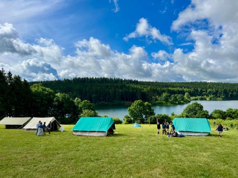 A vibrant lakeside camping scene with tents, people, and lush greenery under a bright blue sky.