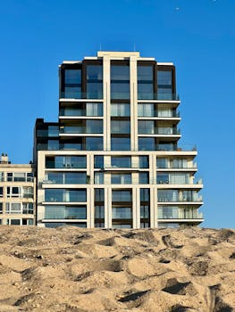 Low angle view of a modern glass facade building, contrasting with sandy foreground under a clear blue sky.