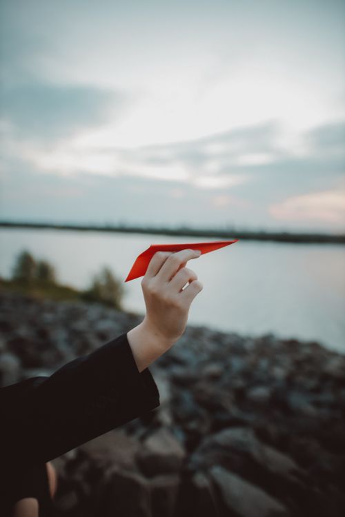 Free A person holds a red paper plane against a blurred lake and sunset sky in Jakarta. Stock Photo
