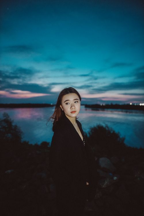 Free Asian woman in black coat stands by a serene lake under a vibrant evening sky. Stock Photo