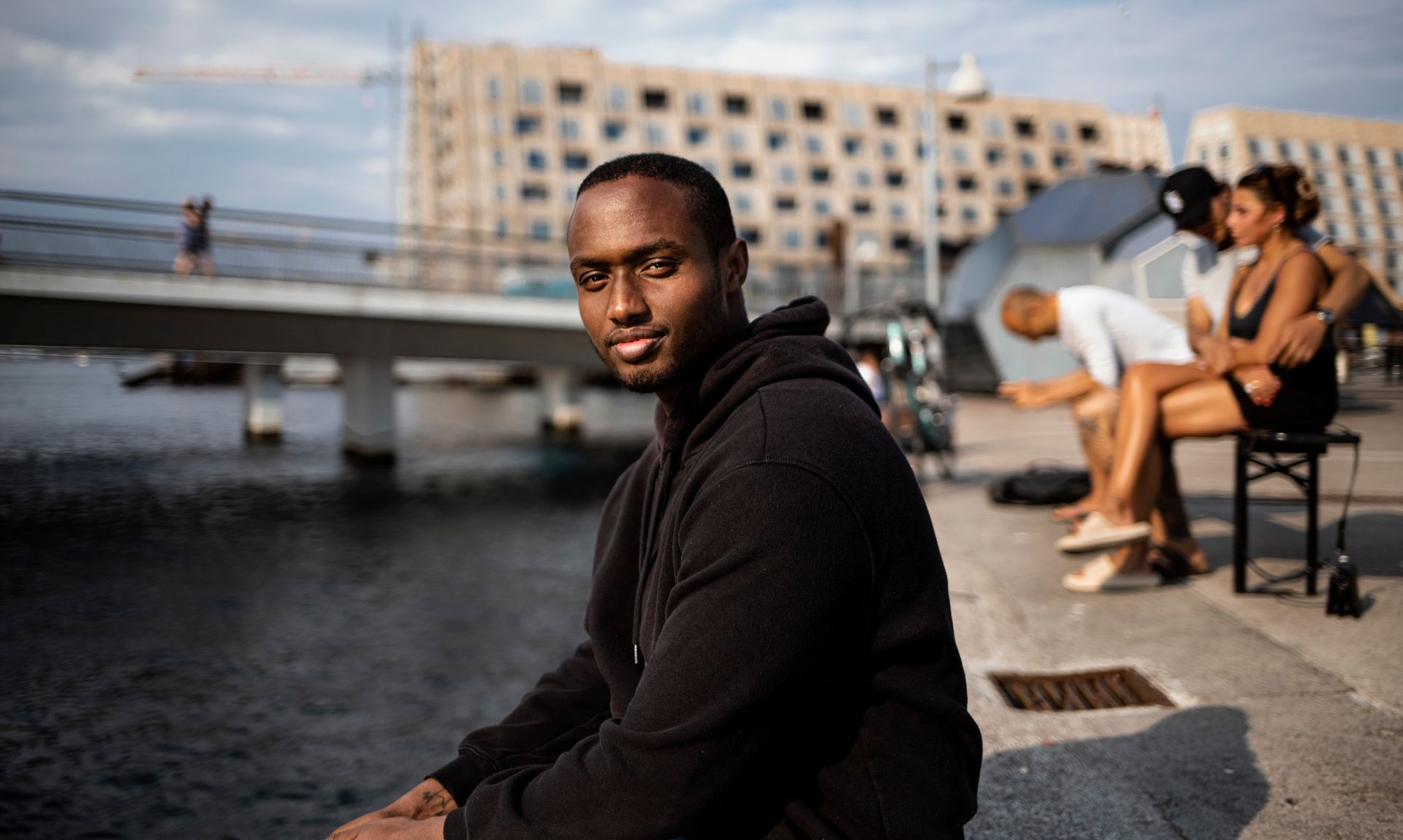 Smiling man in black hoodie sitting by the water in Copenhagen with blurred background.