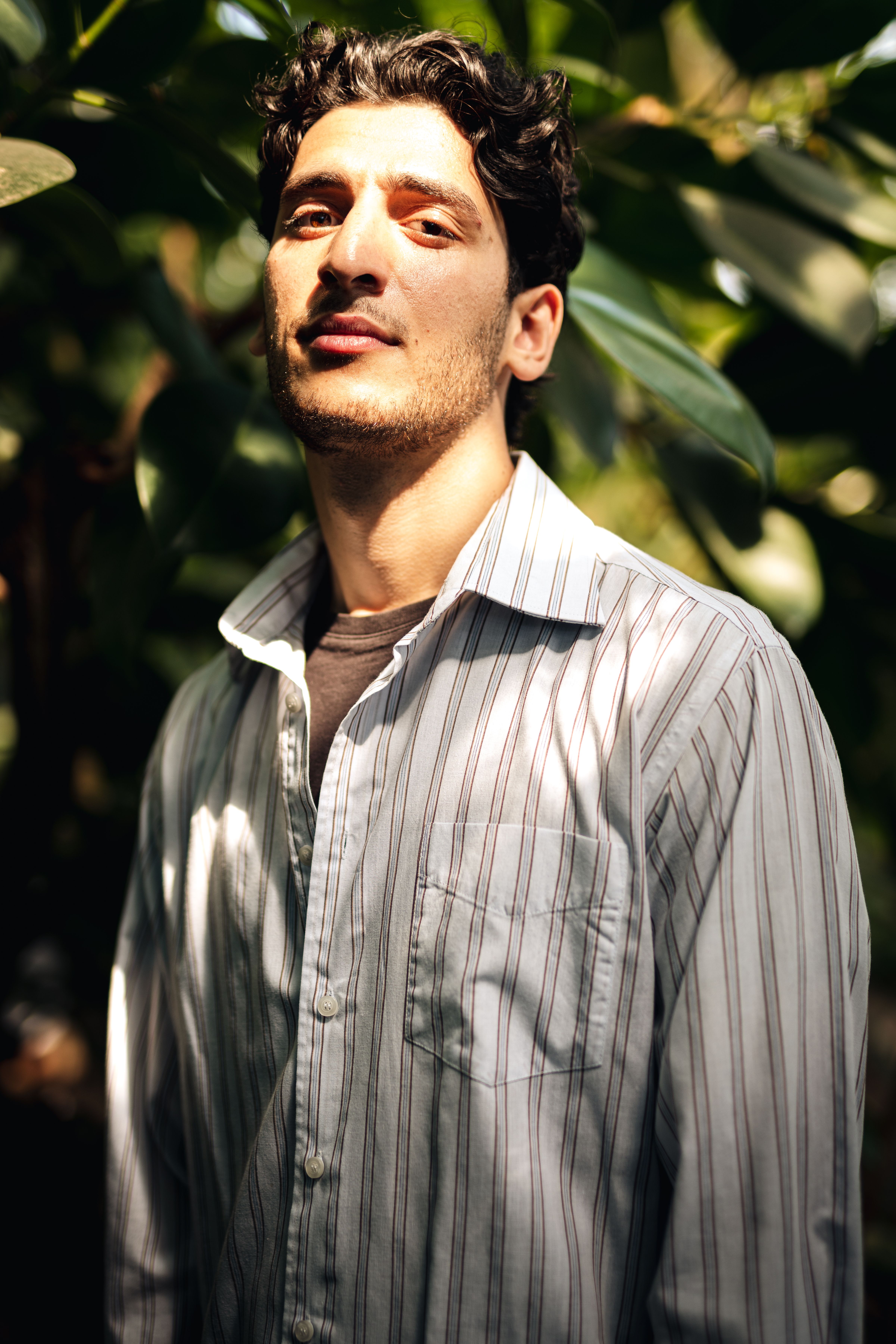 Confident man in striped shirt outdoors with sunlit face, surrounded by green foliage.