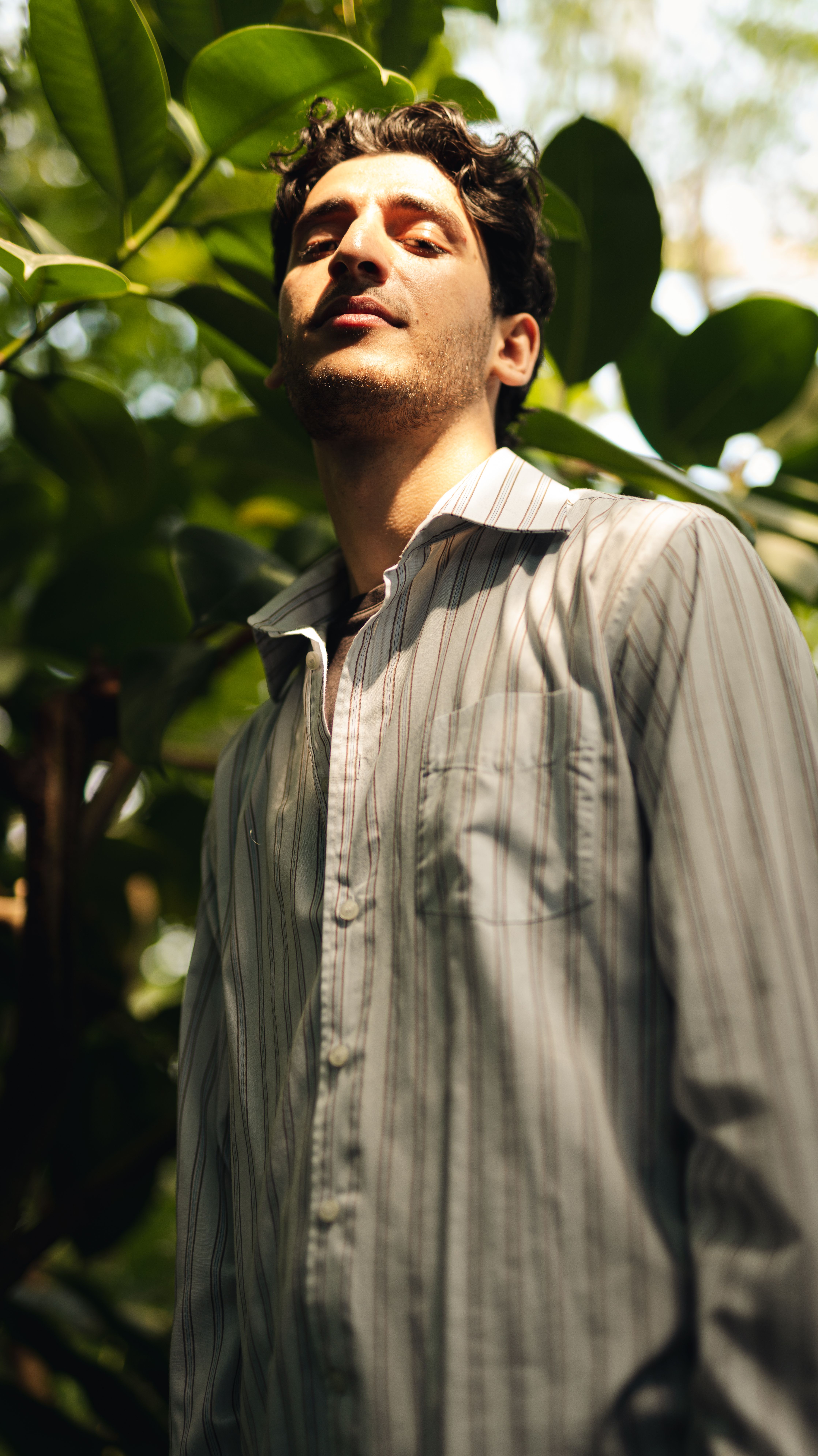 A stylish man in a striped shirt standing outdoors surrounded by lush greenery.