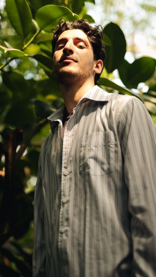 Free A stylish man in a striped shirt standing outdoors surrounded by lush greenery. Stock Photo