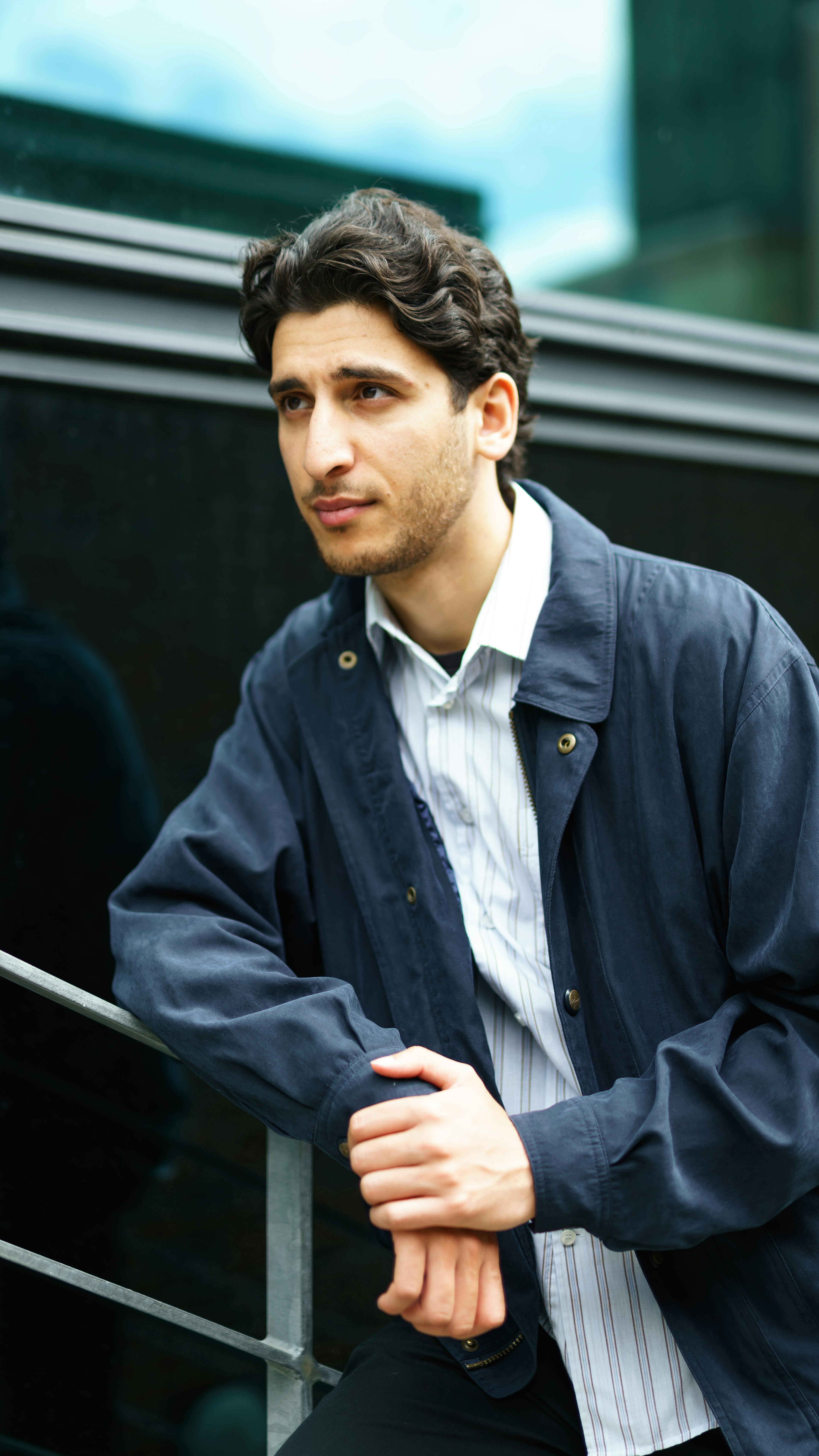 Portrait of a young man in blue jacket leaning on railing in an urban environment.