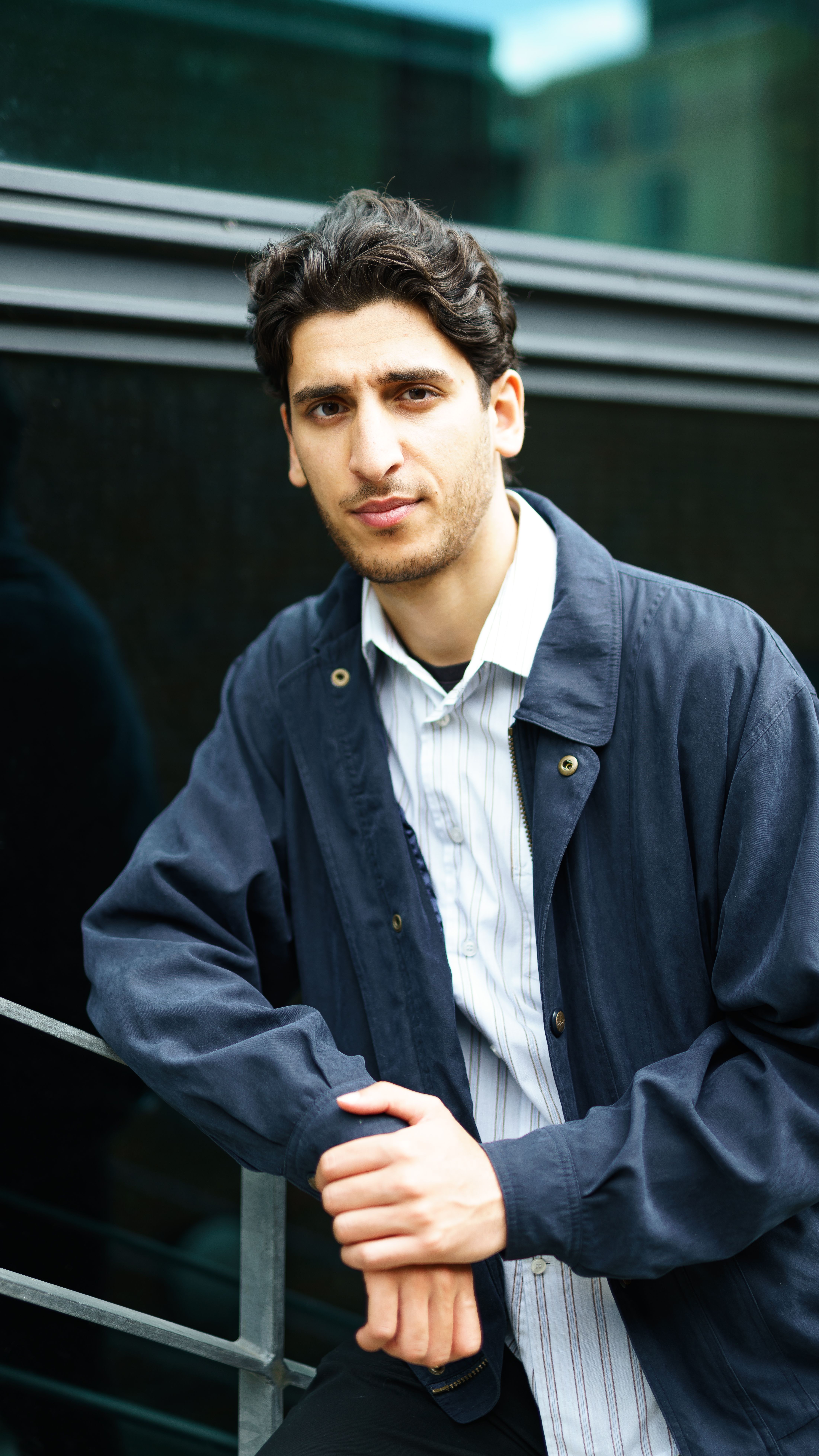 A young man poses confidently in a casual jacket, leaning against a railing outside.