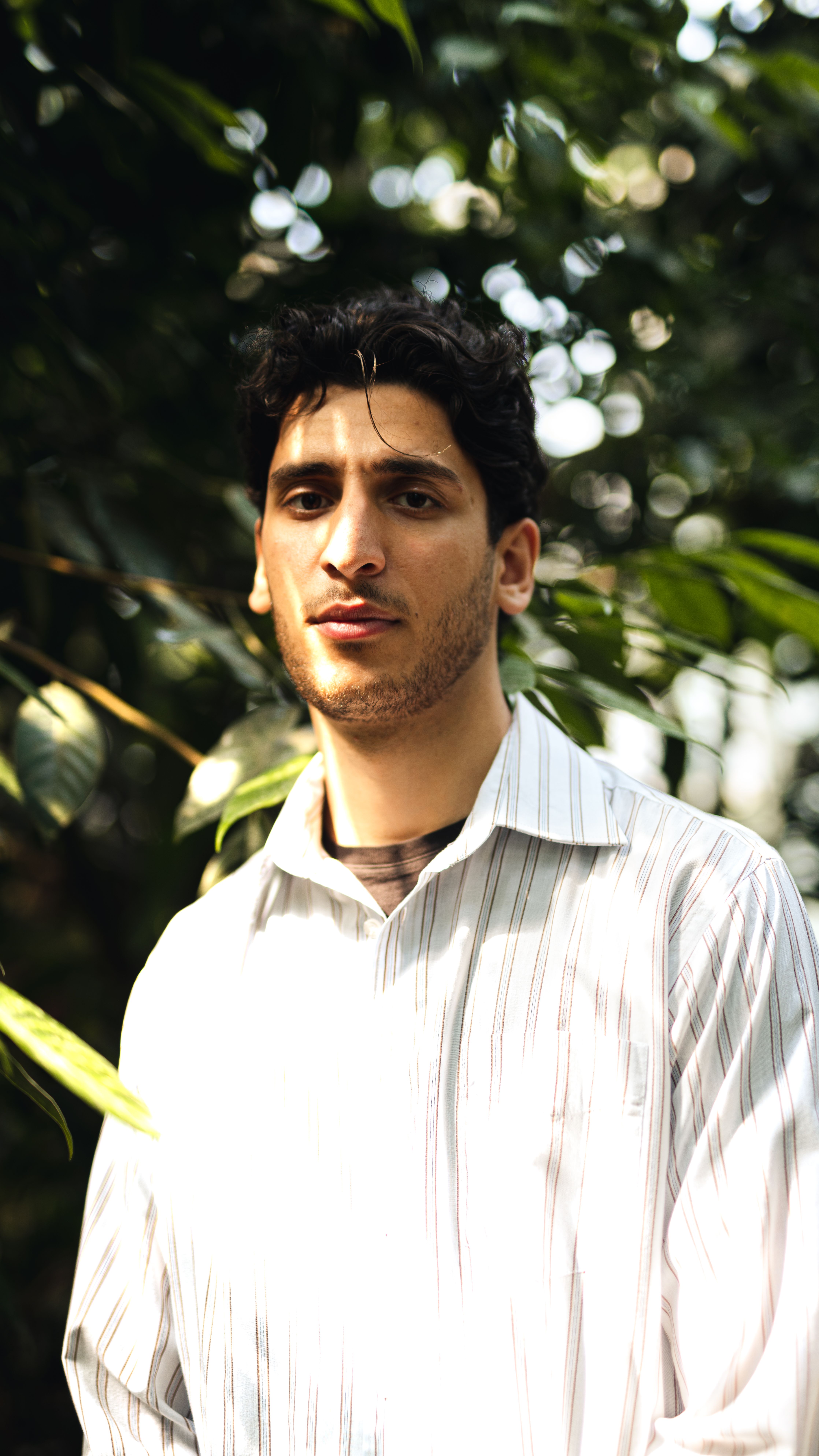 A young man with facial hair wearing a striped shirt in a lush green outdoor environment.