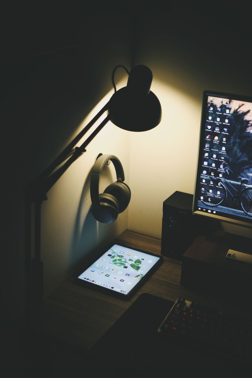 A dimly lit workspace featuring a tablet, table lamp, headphones, and computer screen, creating a cozy atmosphere.