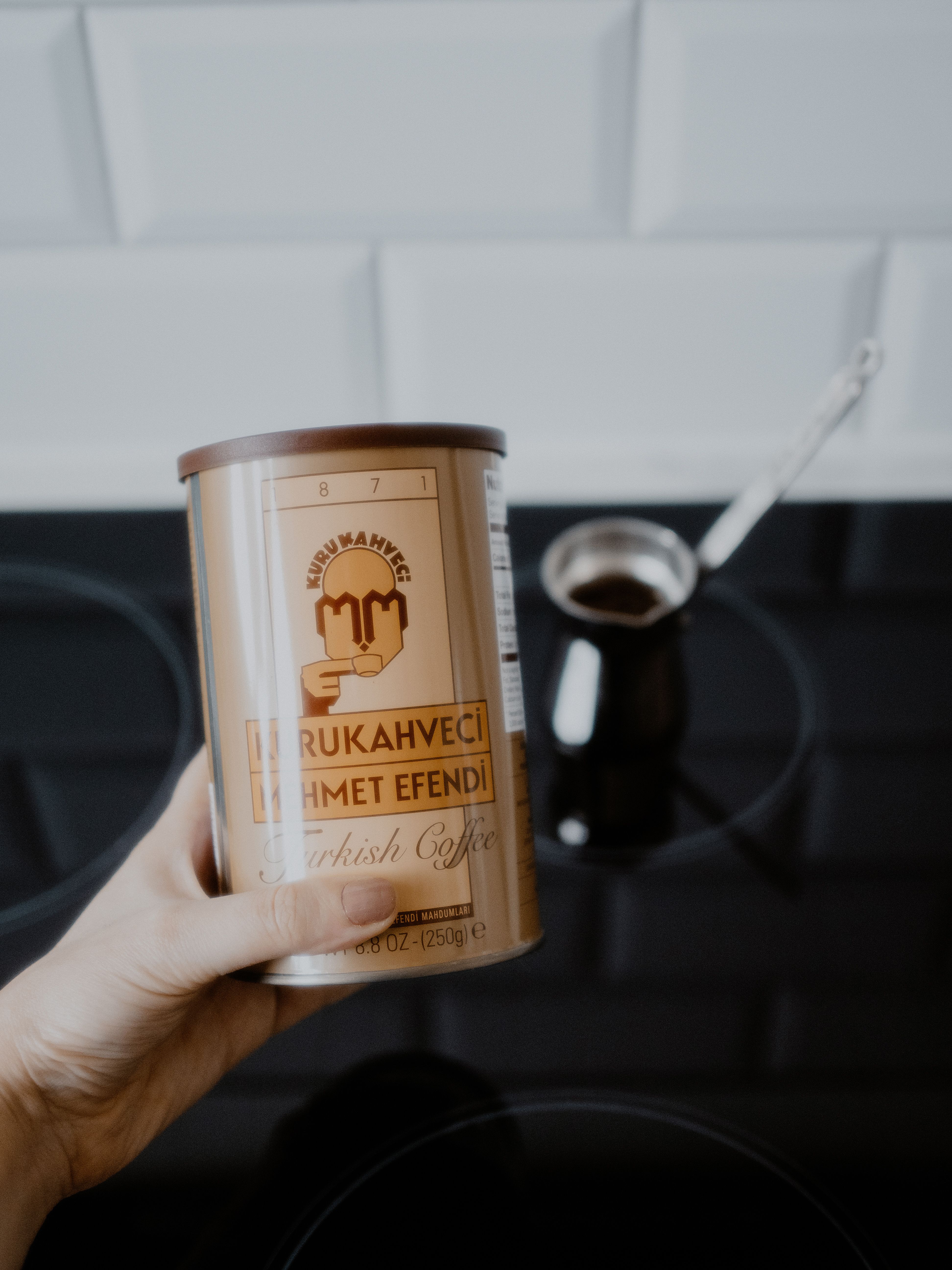 A hand holding Kurukahveci Turkish coffee against a kitchen background, emphasizing traditional brewing.