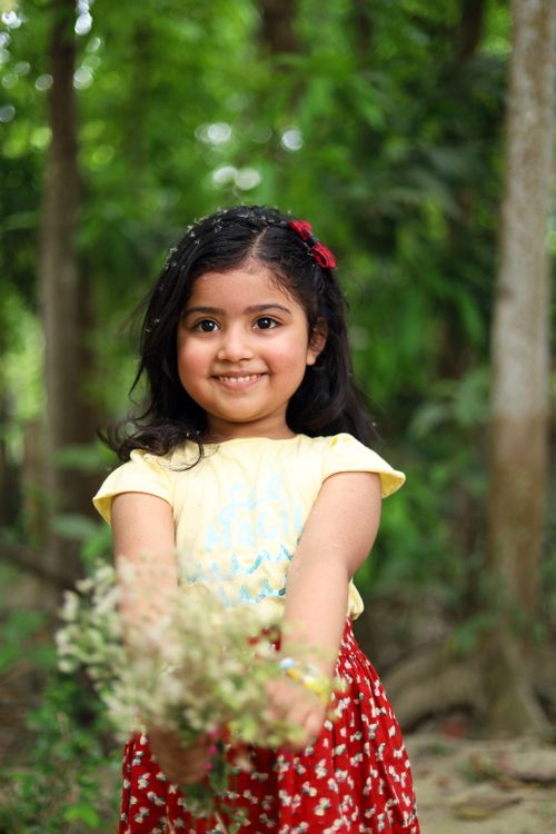 Free Joyful child in a yellow shirt, red skirt, holding flowers outdoors in a green setting. Stock Photo