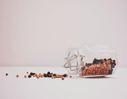 A glass jar tipped over with colorful peppercorns on a minimalist white background.