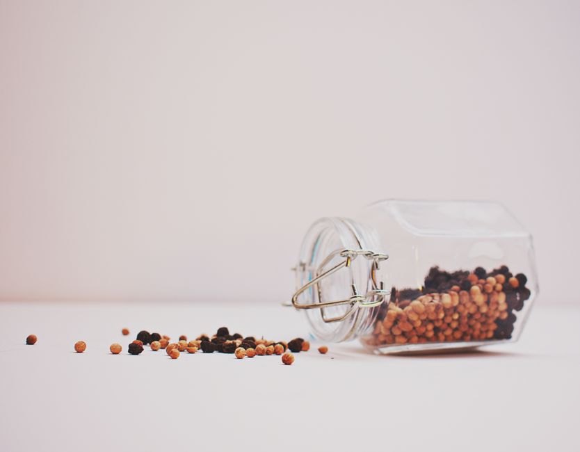 A glass jar tipped over with colorful peppercorns on a minimalist white background.