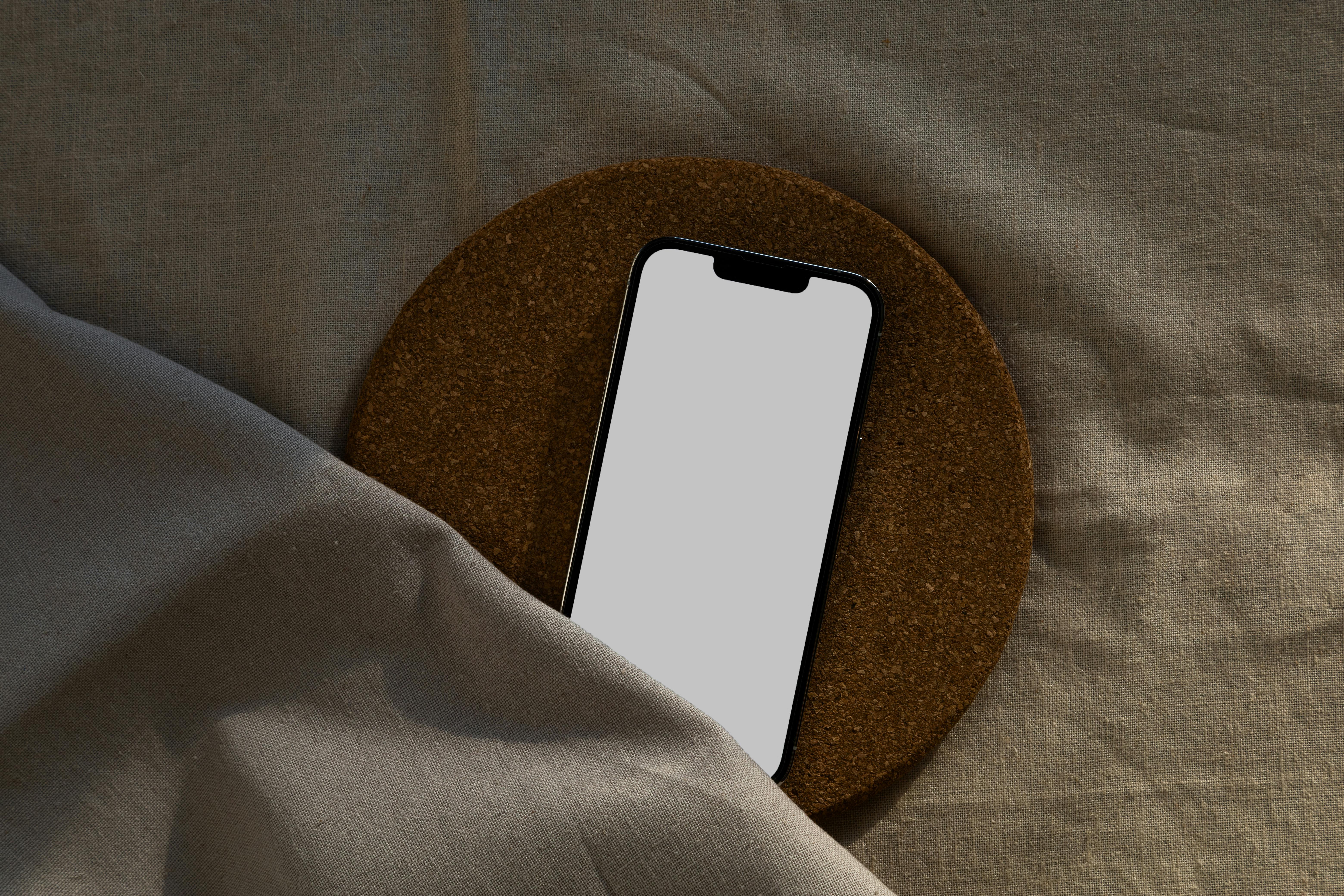 A smartphone with a blank screen on a circular cork surface over a fabric background.