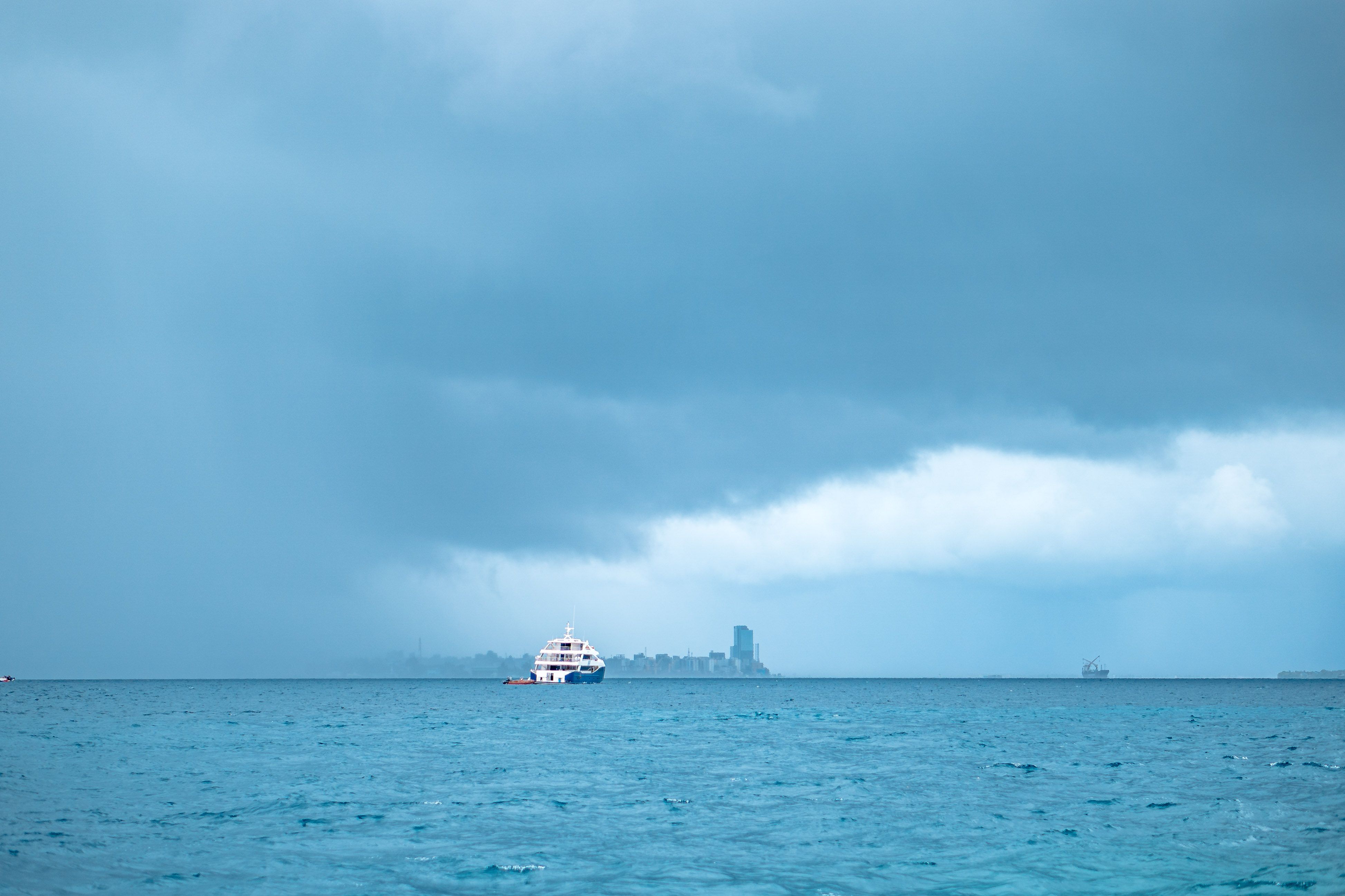 Tranquil seascape featuring a ship with a distant city skyline under dramatic clouds.