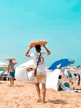 A young vendor carries baked goods on his head on a busy beach, sand beneath his feet.