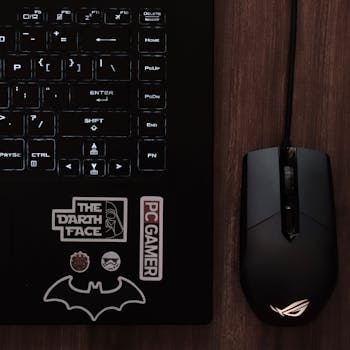 Close-up of a laptop keyboard with a gaming mouse and geek culture stickers on a wooden desk.