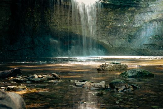 Beautiful waterfall cascading into a serene pool, surrounded by rocks, under a canopy of sunlight.
