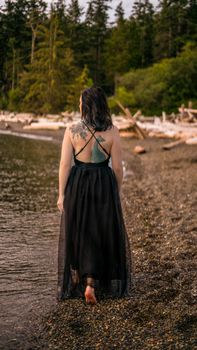 A tattooed woman in a black dress walks along Rosario Beach at sunset, capturing tranquility.