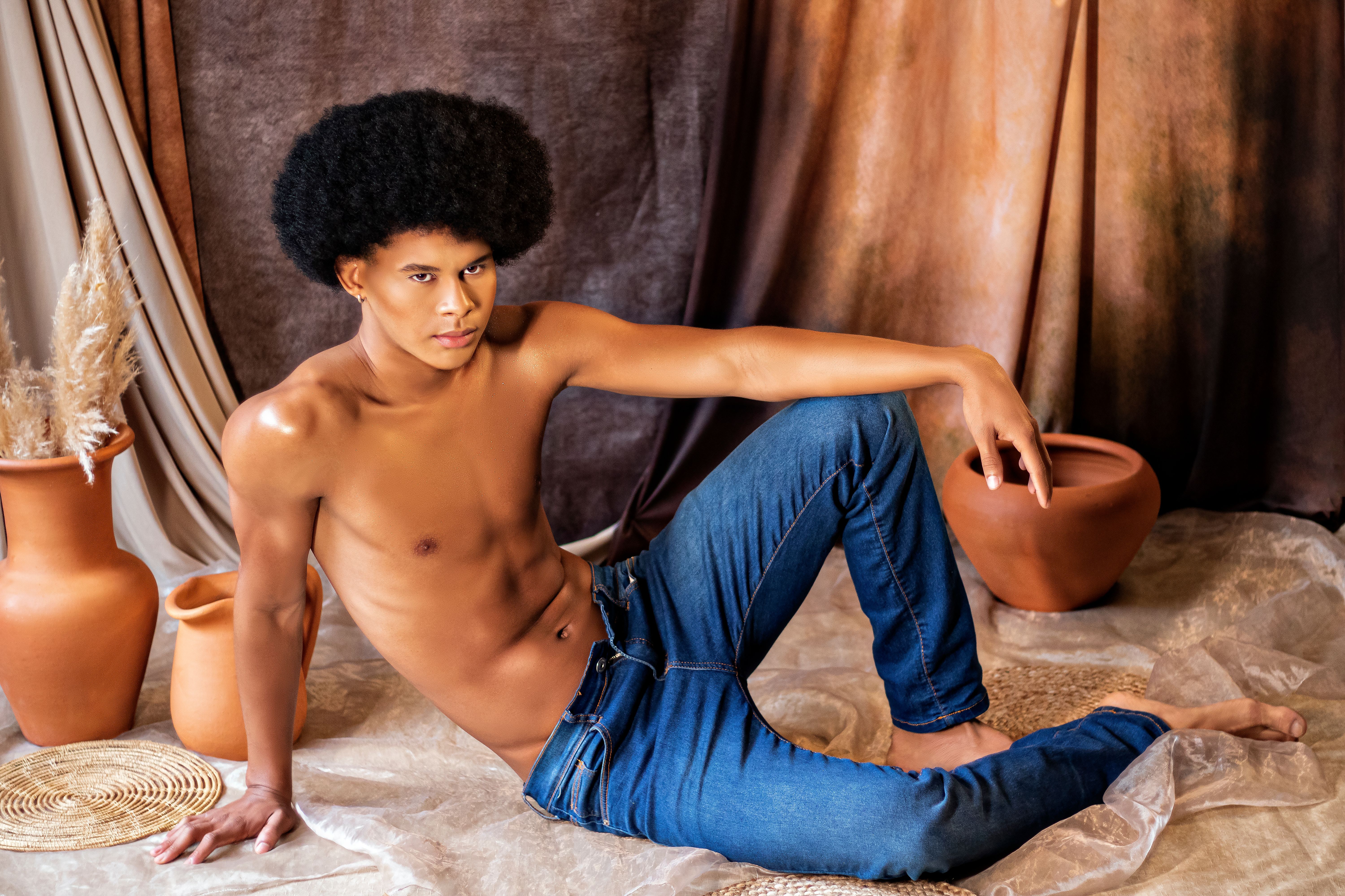 Stylish portrait of an afro-haired man in jeans, posing shirtless indoors against earthy decor.