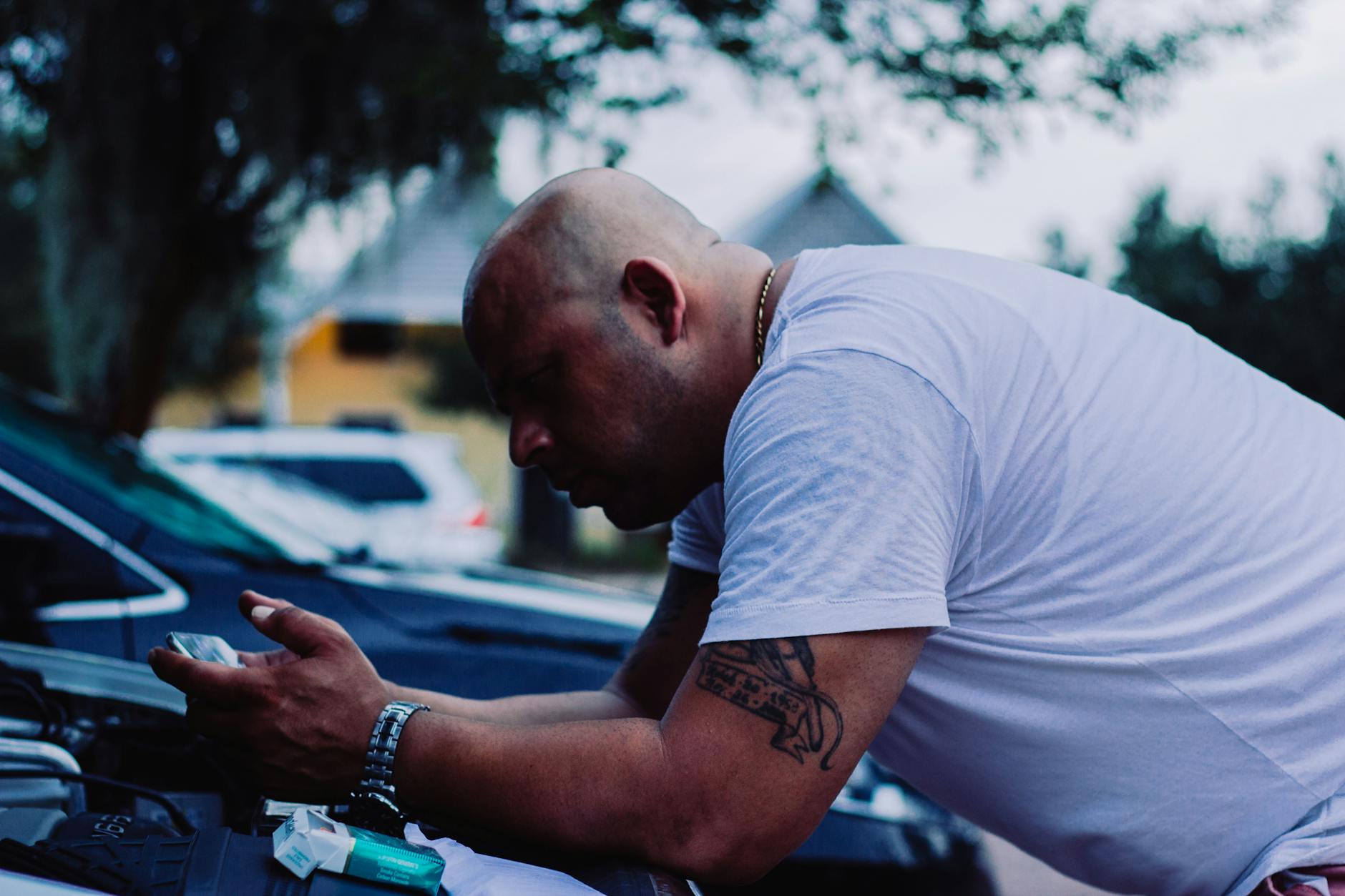 A bald man with tattoos checks a vehicle engine outside. Casual and focused.