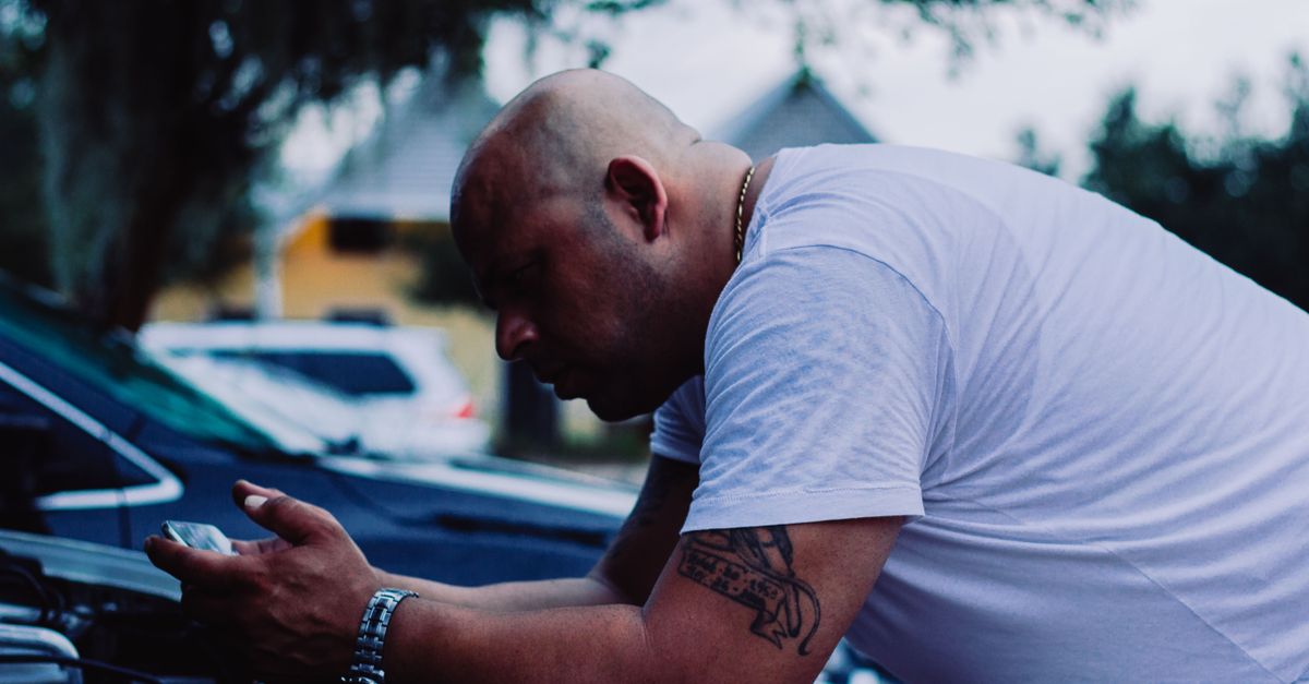 A bald man with tattoos checks a vehicle engine outside. Casual and focused.