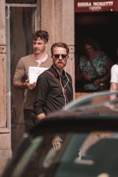 A stylish man in black long sleeves and sunglasses with earphones stands by a car on a sunny street.