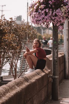 A woman sits by a flower-decorated urban pathway, using a smartphone.