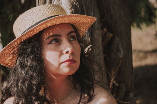Free Portrait of a woman with curly hair and straw hat, gazing up thoughtfully under tree shade. Stock Photo