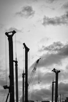 Black and white image of cranes silhouetted against a dramatic cloudy sky, capturing industrial construction.