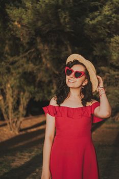Stylish young woman in a red dress with heart-shaped sunglasses, smiling under the sun outdoors.